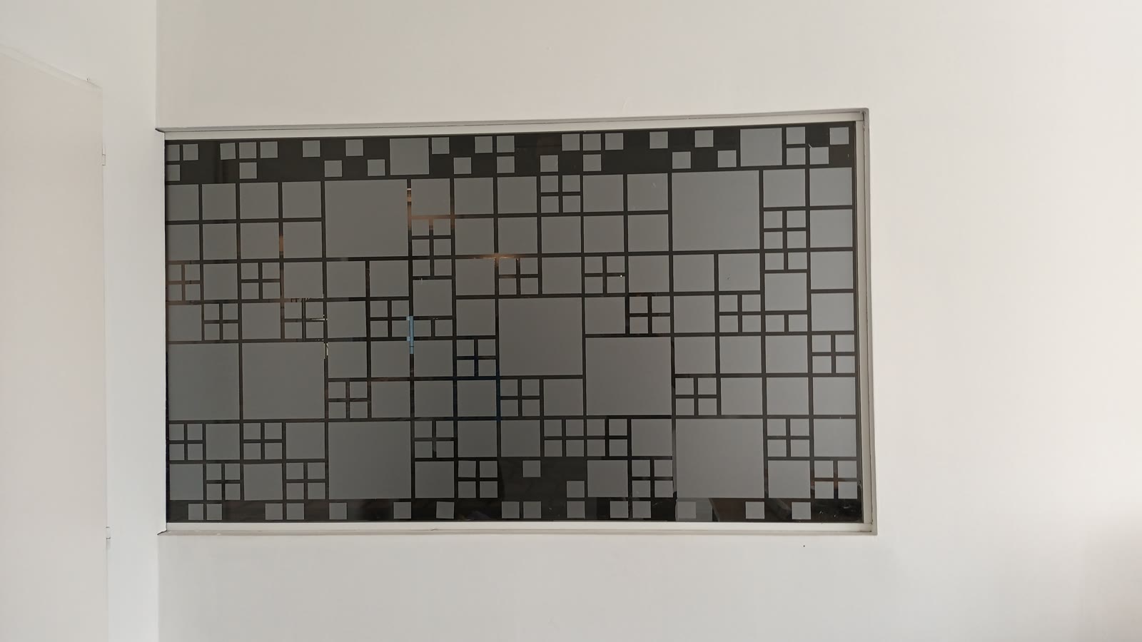 Ventana interior con vinilo esmerilado cortado en cuadrados de distintos tamaños, sobre una pared blanca
